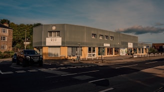 A street scene with a large, rectangular commercial building made of corrugated metal panels and large windows. The building has signage for different businesses and a small parking area in front. A black SUV is parked near the entrance, and a few bollards line the sidewalk. The setting suggests a late afternoon with long shadows and natural light casting a warm tone on the structures.
