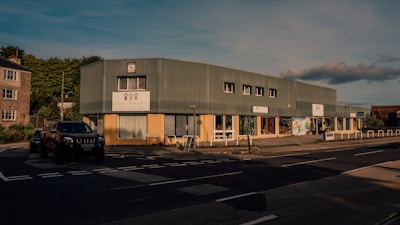 A street scene with a large, rectangular commercial building made of corrugated metal panels and large windows. The building has signage for different businesses and a small parking area in front. A black SUV is parked near the entrance, and a few bollards line the sidewalk. The setting suggests a late afternoon with long shadows and natural light casting a warm tone on the structures.