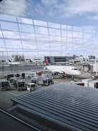 An airport scene features various objects such as a Delta airplane parked at the gate, several luggage carts, and service vehicles. The backdrop includes another aircraft and airport buildings, with a view of the sky through large glass windows reflecting the terminal. Ground crew members are also present, attending to tasks on the tarmac.