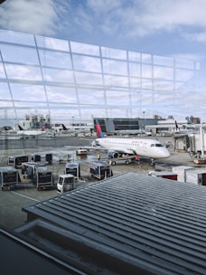An airport scene features various objects such as a Delta airplane parked at the gate, several luggage carts, and service vehicles. The backdrop includes another aircraft and airport buildings, with a view of the sky through large glass windows reflecting the terminal. Ground crew members are also present, attending to tasks on the tarmac.