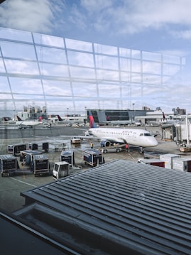 An airport scene features various objects such as a Delta airplane parked at the gate, several luggage carts, and service vehicles. The backdrop includes another aircraft and airport buildings, with a view of the sky through large glass windows reflecting the terminal. Ground crew members are also present, attending to tasks on the tarmac.