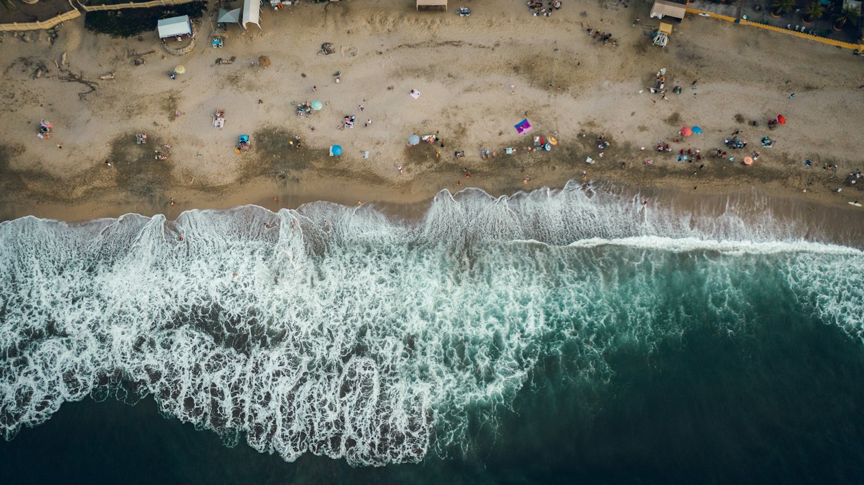 Aerial view of waves breaking white onto a beach with umbrellas and swimmers on the Pacific coast