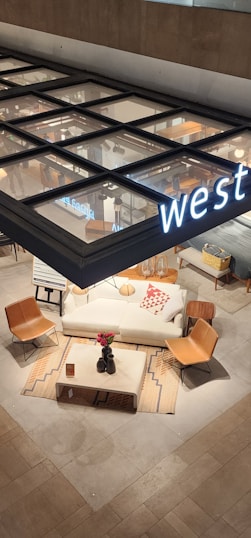 A modern furniture showroom displays a white couch adorned with a decorative red and white pillow. Two brown leather chairs flank the couch, placed on a patterned rug. A square wooden coffee table with a few decorative items sits in the center. The setting is part of a larger showroom with a visible sign above reading 'west'.