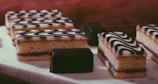 Several rectangular layered pastries topped with a black and white zigzag icing are arranged neatly on a white marble surface. Alongside the pastries, there are a couple of small, dark chocolate cubes. The background has a warm brown hue, creating a cozy, inviting atmosphere.