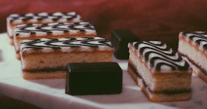 Several rectangular layered pastries topped with a black and white zigzag icing are arranged neatly on a white marble surface. Alongside the pastries, there are a couple of small, dark chocolate cubes. The background has a warm brown hue, creating a cozy, inviting atmosphere.