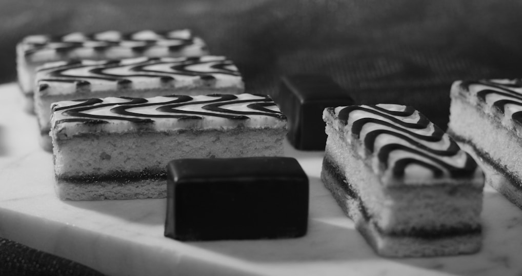 A selection of elegantly arranged desserts, including rectangular pastries with zigzag icing patterns and small chocolate-covered cubes, displayed on a light-colored surface. The image captures a monochromatic aesthetic with a focus on texture and detail.