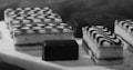 A selection of elegantly arranged desserts, including rectangular pastries with zigzag icing patterns and small chocolate-covered cubes, displayed on a light-colored surface. The image captures a monochromatic aesthetic with a focus on texture and detail.
