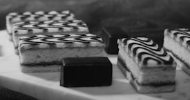 A selection of elegantly arranged desserts, including rectangular pastries with zigzag icing patterns and small chocolate-covered cubes, displayed on a light-colored surface. The image captures a monochromatic aesthetic with a focus on texture and detail.