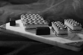 Rectangular pastries with intricate black and white swirl patterns are arranged on a marble serving board. The pastries are accompanied by small, smooth chocolate pieces, all placed on a dark fabric background.