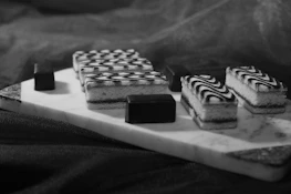 Rectangular pastries with intricate black and white swirl patterns are arranged on a marble serving board. The pastries are accompanied by small, smooth chocolate pieces, all placed on a dark fabric background.