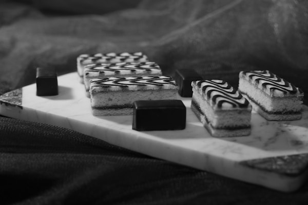 Rectangular pastries with intricate black and white swirl patterns are arranged on a marble serving board. The pastries are accompanied by small, smooth chocolate pieces, all placed on a dark fabric background.