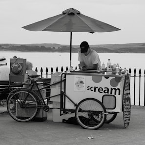 A monochrome image of an ice cream vendor with a cart on a seafront promenade. The vendor leans on the cart, looking at a phone, beneath a large umbrella. The cart has wheels and a sign that reads 'scream', with various bottles and containers placed on top. Behind, there is a fence and a view of a body of water with distant hills.