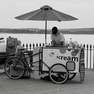 A monochrome image of an ice cream vendor with a cart on a seafront promenade. The vendor leans on the cart, looking at a phone, beneath a large umbrella. The cart has wheels and a sign that reads 'scream', with various bottles and containers placed on top. Behind, there is a fence and a view of a body of water with distant hills.