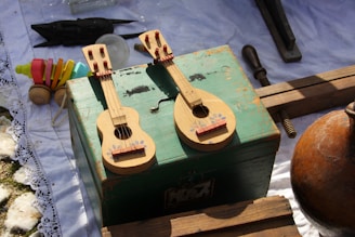 Two small string instruments resembling miniature guitars rest on an aged, green wooden box. The instruments feature painted floral decorations below their strings. Surrounding them are a collection of old tools and toys, including colorful toy blocks and vintage wooden pieces, all placed on a white cloth with lace edges.