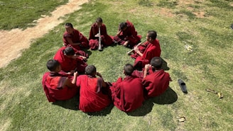A group of young monks dressed in red robes sit in a circle on green grass. One of them is playing a wind instrument, while the others attentively listen or chat among themselves. The scene is set outdoors on a sunny day, indicated by the shadows and bright lighting.