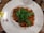 A plate featuring a serving of fusilli pasta covered in a red tomato-based sauce, topped with fresh green arugula. The dish is placed on a white, slightly speckled plate, which has a branded logo and a yellow 'gluten' label at the edge. The table surface is wooden, and there are specks of sauce on the plate's rim.