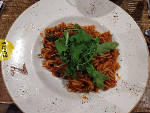 A plate featuring a serving of fusilli pasta covered in a red tomato-based sauce, topped with fresh green arugula. The dish is placed on a white, slightly speckled plate, which has a branded logo and a yellow 'gluten' label at the edge. The table surface is wooden, and there are specks of sauce on the plate's rim.