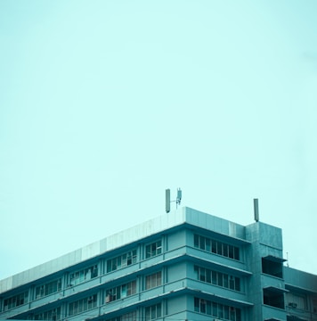A multi-story building with a flat roof is captured against a clear blue sky. The structure features several rows of windows and has a few communication antennas installed on the rooftop.