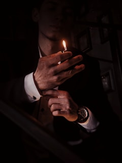 A person is illuminated by the small flame of a lighter in a dimly lit environment. The focus is on the hands and lighter, with the glow casting light onto a wristwatch. The background is dark, adding to the dramatic effect and highlighting the person's formal attire.