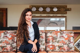 A woman with long, wavy hair wearing a navy blue blazer and sitting comfortably on a sofa. Behind her are five clocks mounted on a wooden panel, showing different world time zones such as London, Paris, São Paulo, New York, and Tokyo. The sofa has a textured pattern in orange, white, and green.