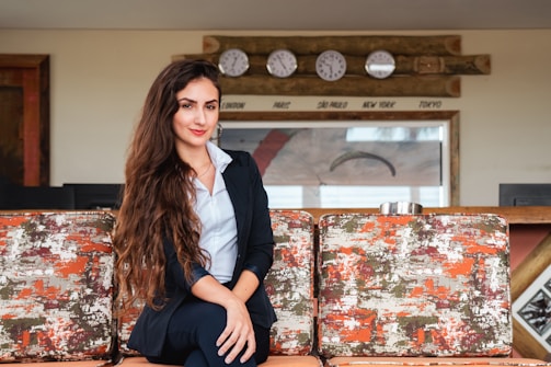 A woman with long, wavy hair wearing a navy blue blazer and sitting comfortably on a sofa. Behind her are five clocks mounted on a wooden panel, showing different world time zones such as London, Paris, São Paulo, New York, and Tokyo. The sofa has a textured pattern in orange, white, and green.