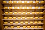 Several neatly arranged wheels of cheese rest on multiple wooden shelves, creating a symmetrical and organized display. The cheeses are uniform in shape and have a smooth, glossy surface, suggesting they are well-cared for.