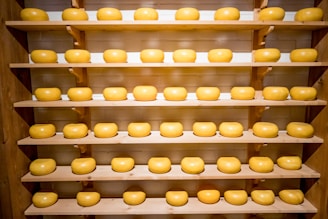 Several neatly arranged wheels of cheese rest on multiple wooden shelves, creating a symmetrical and organized display. The cheeses are uniform in shape and have a smooth, glossy surface, suggesting they are well-cared for.