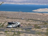 A recreational vehicle is driving along a winding road in a desert landscape, with brown and sparse vegetation. In the distance, a large blue lake is visible, surrounded by arid hills and mountains under a clear sky.
