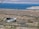 A recreational vehicle is driving along a winding road in a desert landscape, with brown and sparse vegetation. In the distance, a large blue lake is visible, surrounded by arid hills and mountains under a clear sky.