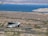 A recreational vehicle is driving along a winding road in a desert landscape, with brown and sparse vegetation. In the distance, a large blue lake is visible, surrounded by arid hills and mountains under a clear sky.