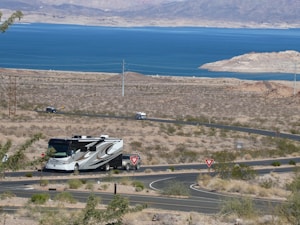 A recreational vehicle is driving along a winding road in a desert landscape, with brown and sparse vegetation. In the distance, a large blue lake is visible, surrounded by arid hills and mountains under a clear sky.