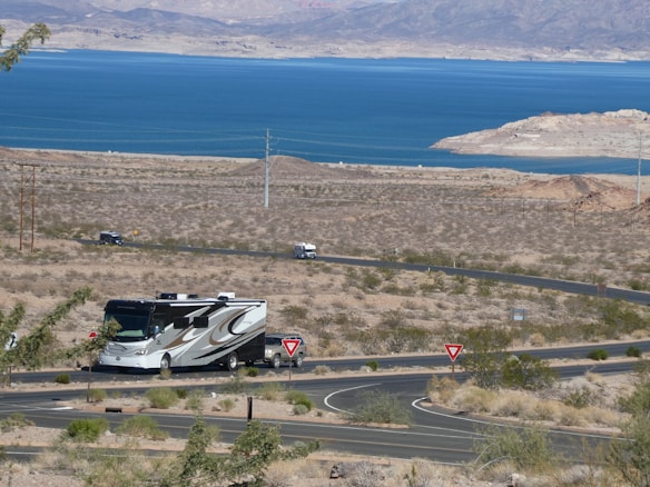 A recreational vehicle is driving along a winding road in a desert landscape, with brown and sparse vegetation. In the distance, a large blue lake is visible, surrounded by arid hills and mountains under a clear sky.