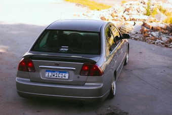 A silver car is parked on a concrete surface near a pile of rubble and debris. The vehicle has a visible Brazilian license plate, and stickers are on the rear window. The setting appears to be an outdoor area with bright sunlight casting shadows.