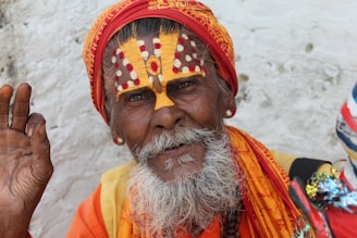 An elderly man with a long white beard and distinct facial paint is wearing a bright orange and red turban. The intricate design on his face has white and red dots with a bold yellow pattern. His hand is lifted in a gesture, possibly a blessing or greeting. He is adorned in colorful clothing with vibrant hues.