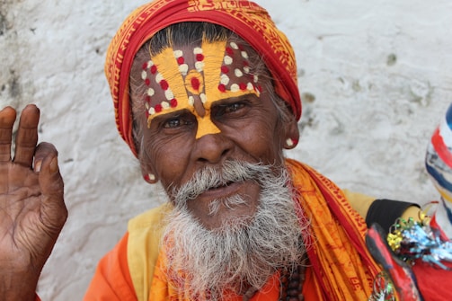 An elderly man with a long white beard and distinct facial paint is wearing a bright orange and red turban. The intricate design on his face has white and red dots with a bold yellow pattern. His hand is lifted in a gesture, possibly a blessing or greeting. He is adorned in colorful clothing with vibrant hues.