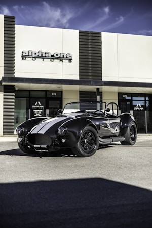 A classic black sports car with white racing stripes is parked in front of a modern building with the sign 'alpha one motors'. The car features a sleek, aerodynamic design with prominent headlights and a convertible top. The building has a minimalist style with large glass windows.
