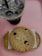 A large chocolate chip cookie is placed on a wooden tray with a napkin underneath. Next to it is a tall plastic cup filled with iced coffee, topped with bubbles and a green straw. The setting is on a pink surface.