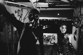 Two people are standing in a dimly lit, graffiti-covered room. One is wearing a gas mask and dark clothing, while the other is dressed in a leather outfit with a futuristic look. There is a cityscape visible in the background through a window or opening.
