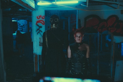Two individuals stand in a dimly lit, graffiti-covered building. The person on the left wears a dark outfit with chains and a gas mask. The person on the right, with red hair styled in pigtails, is dressed in a black leather outfit with gloves and a choker. The environment is slightly eerie and cluttered.