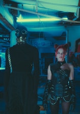 A dimly lit, futuristic setting with two individuals in elaborate costumes. One person is wearing a long black outfit and a gas mask, giving an impression of anonymity. The other person, with vivid red hair styled in buns, is dressed in intricate black leather attire with belts and gloves, exuding a punk or cyber-goth aesthetic. Neon blue lighting creates an industrial and otherworldly atmosphere.