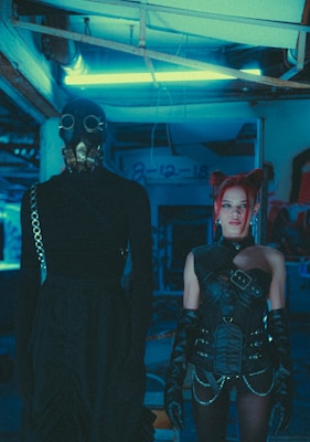A dimly lit, futuristic setting with two individuals in elaborate costumes. One person is wearing a long black outfit and a gas mask, giving an impression of anonymity. The other person, with vivid red hair styled in buns, is dressed in intricate black leather attire with belts and gloves, exuding a punk or cyber-goth aesthetic. Neon blue lighting creates an industrial and otherworldly atmosphere.
