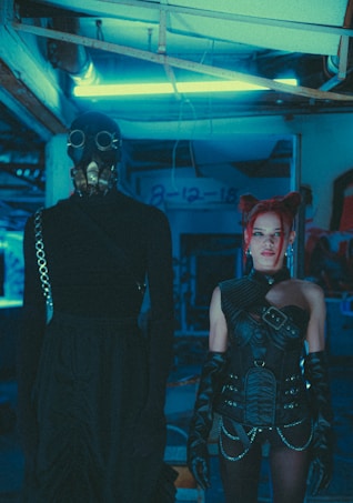 A dimly lit, futuristic setting with two individuals in elaborate costumes. One person is wearing a long black outfit and a gas mask, giving an impression of anonymity. The other person, with vivid red hair styled in buns, is dressed in intricate black leather attire with belts and gloves, exuding a punk or cyber-goth aesthetic. Neon blue lighting creates an industrial and otherworldly atmosphere.
