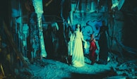 A dimly-lit, eerie setting with figures dressed in extravagant and dark clothing. The background features graffiti on a wall, with exposed beams and rubble on the ground, creating a haunting atmosphere.