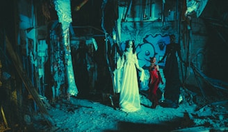 A dimly-lit, eerie setting with figures dressed in extravagant and dark clothing. The background features graffiti on a wall, with exposed beams and rubble on the ground, creating a haunting atmosphere.