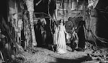 In a dilapidated and graffiti-covered space, three figures are dressed in elaborate costumes. The central figure wears a long, flowing white dress with a painted face, evoking a ghostly appearance. On either side, two figures are dressed in dark, theatrical outfits, adding to a mysterious and eerie atmosphere. The environment is cluttered, with debris and tattered materials hanging from the ceiling.