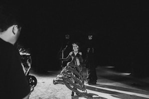 In a dark environment, two individuals are dressed in elaborate costumes with dark and gothic elements. One person is at the center wearing a corset and a skirt that appears to be made of thick, snake-like coils. Two figures in the background wear masks and dark clothing. A person on the left is holding a camera, suggesting a photoshoot or film set.