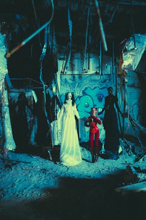 A dimly lit, eerie scene in an abandoned, graffiti-covered space. Two figures are prominently displayed: one in a long, flowing white gown with a ghostly presence, and another dressed in red with a devilish appearance. Surrounded by hanging debris and dark shadows, the setting evokes a sense of decay and abandonment.