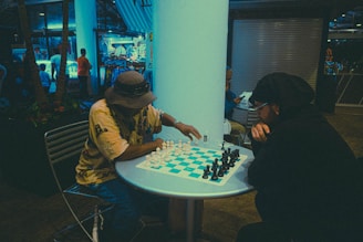 Two people are engaged in a game of chess at a small round table in a dimly lit indoor setting, surrounded by a backdrop of shops or stalls. One player is wearing a hat and a light-colored shirt, while the other is dressed in dark clothing.