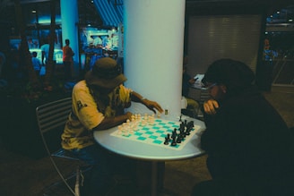 Two people are engaged in a chess game at a small round table. One player is wearing a hat and a yellow shirt, while the other is dressed in dark clothing. The setting appears to be indoors or in a covered outdoor area with a relaxed, casual atmosphere. In the background, there are visible shops with blue lighting and people walking around, giving a sense of an urban or commercial environment.