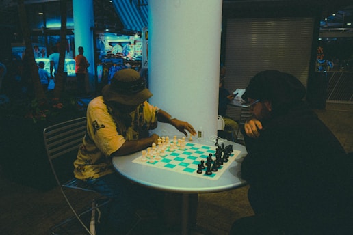 Two people are engaged in a chess game at a small round table. One player is wearing a hat and a yellow shirt, while the other is dressed in dark clothing. The setting appears to be indoors or in a covered outdoor area with a relaxed, casual atmosphere. In the background, there are visible shops with blue lighting and people walking around, giving a sense of an urban or commercial environment.